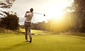 A golfer swings a club on a well-maintained golf course during sunset, surrounded by trees and bathed in golden sunlight.