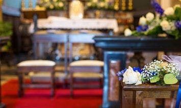 Bouquet of flowers rests on a wooden bench. The setting appears to be a church interior with an altar adorned with flowers, candles, and a red carpet leading to the front.