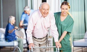 An elderly man using a walker, assisted by a smiling caregiver in green scrubs, is in the foreground. In the background, another elderly individual with a walker is attended to by another caregiver.