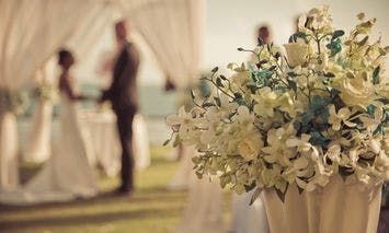 A bouquet of white and blue flowers sits in the foreground; in the background, a couple exchanges vows under a white draped canopy in an outdoor setting.